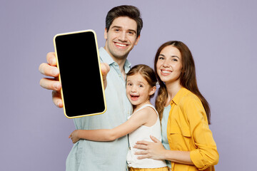 Wall Mural - Young parents mom dad with child kid girl 6 years old wearing blue yellow casual clothes hold use show close up blank screen mobile cell phone isolated on plain purple background. Family day concept.