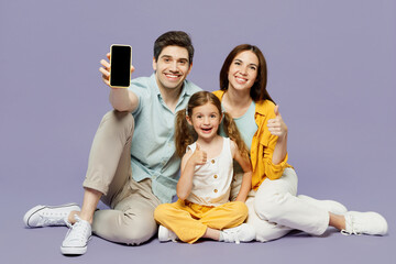 Wall Mural - Full body young parents mom dad with child kid daughter girl 6 years old wear yellow casual clothes sit hold use blaank screen mobile cell phone isolated on plain purple background Family day concept