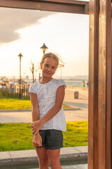 a cute little girl stands on the background of the sea and the playground. smiling and looking ahead