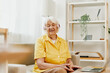 © SHOTPRIME STUDIO - An elderly woman wearing headphones with a laptop sits on the couch at home and works in a yellow shirt in front of a window.