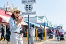Cheerful Asian Chinese Female Tourist Searching For Best Seafood Restaurant On Phone Near Route 66 End Of The Trail Sign During Spring Holiday At Santa Monica Pier In The Us
