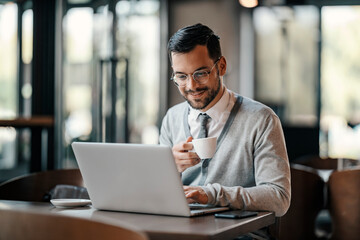 A happy young trendy businessman in smart causal is sitting in cafe, drinking coffee and typing on a laptop.