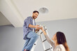 © Studio Romantic - Smiling man changing new electric bulb at home. His wife helping her husband mounting ceiling lamp. Cheerful man standing on ladder holding new LED light bulb, low angle view