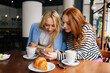 © dikushin - Portrait of two happy young women sitting at table with coffee at cafe by window, having conversation and showing each other something on mobile phones. Concept of woman friendship