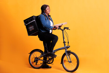 Wall Mural - African american pizzeria courier carrying pizza carton box delivering takeaway order to client during lunch time, using bike as transportation in studio. Take out food service and concept