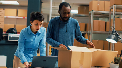 Team of entrepreneurs packing order for shipment, using cardboard boxes and warehouse stock. Cheerful people working on supply chain and distribution logistics, quality control. Handheld shot.