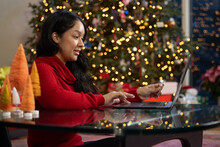 Hispanic Woman Holding Credit Card In Her Left Hand While Online Shopping On Her Computer In Front Of Her Christmas Tree At Home