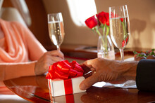 Detail Shot Of Mature Couple Sitting Across From One Another At Table With Champagne Glasses Aboard A Private Airplane, Man Holding A Wrapped Gift.  