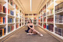 Portrait Of Young Female Ballerina Sitting On Floor Of Library Pulling Looking Off Camera 