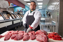 Portrait Of Butcher Standing Tall Wearing Hair Net, Apron And Gloves Behind Counter Of Meat Market Of Grocery Store Making Cuts Of Steak 