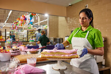 Female Baker Spreading On Strawberry Frosting To Build Layer Cake In Latin Market Bakery, Baker In Background Building Birthday Cake 