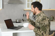 Happy soldier with cup of drink using video chat on laptop at white marble table in kitchen. Military service