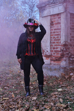 A woman in the guise of Baron Sabbath poses in red smoke. The model is dressed in a corset, a cardigan and a top hat decorated with skeleton figures. Model looking at the camera