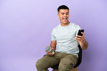 Wall Mural - Young man sitting on a chair over isolated background holding coffee to take away and a mobile