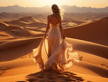Scenic Back Portrait Of A Beautiful Young Woman Model In A Long White Silk Dress Walking On Sand Dunes Crest In The Desert Leaving Footprints Trail On A Sand.