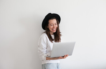 Wall Mural - Young millennial woman wearing black hat using laptop, working online, white background. Copy space left and right.