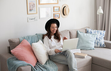 Wall Mural - Young woman using laptop, working online, wearing black hat, smiling, sitting on a comfy couch. Trendy hipster cozy interior. Artist living slow freelance life. Millennial lifestyle. 