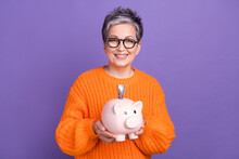 Photo Of Positive Intelligent Woman With Short Haircut Dressed Orange Sweater Hold Pig Money Box Isolated On Purple Color Background