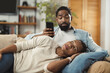 © Goran - Young black woman lying on her husband's lap and wondering to whom is he sending a text message while relaxing together on sofa