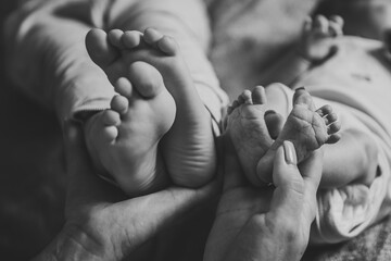 Baby feet in mom hands. Mother holding child's legs. Little infant lying on bed at home. Mom hold son legs. Closeup. Tiny, bare feet of newborn baby girl and boy. Sleep daughter. Black white photo