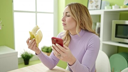 Wall Mural - Young beautiful hispanic woman eating banana using smartphone at dinning room