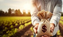 A Hand Holds Out A Dollar Money Bag On A Background Of A Farm Field. Lending Farmers And Agricultural Enterprises For Purchase Land And Seed Material, Equipment Modernization Support And Subsidies.