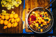 A Pan Of Fruit Cobbler Being Prepared On A Wood Cutting Board Next To Sliced Peaches And Pineapple At Night, Closeup