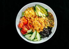 A White Bowl With Chicken, Avocado, Tomatoes, Beans And Other Grains And Vegetables Against A Black Background