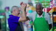 © Marco - Happy diverse senior employees celebrating success with high-five standing in supermarket aisle. Caucasian manager engaging with workforce teamwork with African American colleague