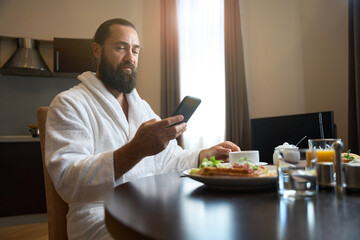 Man in bathrobe sits at table with phone and breakfast