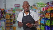 © Marco - Joyful Happy Brazilian senior employee of supermarkert wearing apron and holding tablet device, portrait of a black older male staff depicting job occupation at retail store