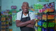 © Marco - Portrait of a joyful African American senior employee of supermarket wearing apron and smiling at camera inside grocery store aisle and arms crossed