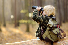Little Boy Scout With Binoculars During Hiking In Autumn Forest. Child Is Sitting On Large Fallen Tree And Looking Through A Binoculars.