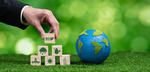 Businessman hand holding wooden cube with eco symbol and paper globe on fertile soil background. Environmental awareness and sustainable energy. Clean and renewable energy for greener ecology. Alter