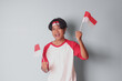 Portrait of attractive Asian man in t-shirt with red and white ribbon on head, holding indonesia flag while raising his fist, celebrating success. Isolated image on gray background