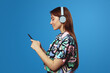 Side view of young student girl using mobile phone and looking at screen, wearing headphones while listening to music, isolated on blue background. People, lifestyle and emotion concept