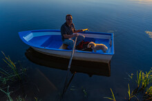 A 60-year-old Caucasian Man On A Blue Wooden Boat With Oars In His Hands With A Poodle Dog Lit By The Sun At Sunset.