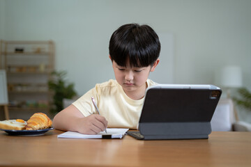 Attentive boy writing in notebook while sitting at desk and doing homework.