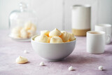 Small meringues in a white bowl over light pink and white wooden background, two cups of coffee, creamer behind