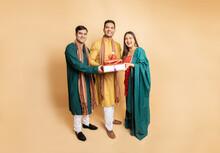 Happy Young Indian Family Brother Sister Wearing Traditional Cloths Holding Red Gift Boxes Standing Isolated On Beige Background. Celebrating Diwali Festival.