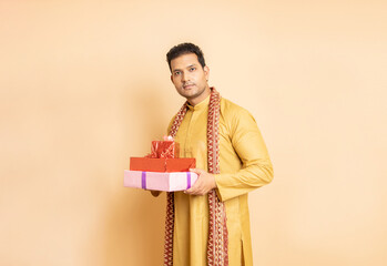 Wall Mural - Happy young handsome indian man wearing traditional cloths holding red gift boxes celebrating of diwali festival isolated on beige background.