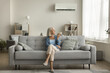 © fizkes - Positive relaxed senior lady using remote control for turning AC on, starting cooling domestic equipment, relaxing on comfortable home couch in cozy modern apartment interior