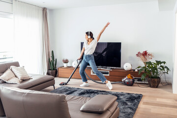 Wall Mural - Young happy woman listening and dancing to music while cleaning the living room floor with a vaccum cleaner