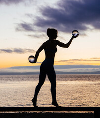 Silhouette of young woman doing workout on a bench with crystal ball(s), in front of ocean at sunrise or sunset