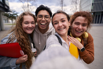 Wall Mural - Group of four students having fun by taking a selfie outdoors. The photo is taken outside the campus of the university. They look very happy and look at the camera.