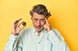 © Asier - Man holding a physical bitcoin, yellow backdrop showing a disappointment gesture with forefinger.