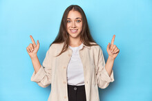 Stylish Young Woman In An Overshirt On A Blue Background Indicates With Both Fore Fingers Up Showing A Blank Space.