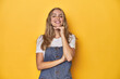 © Asier - Young blonde Caucasian woman in denim overalls posing on a yellow background, smiling happy and confident, touching chin with hand.