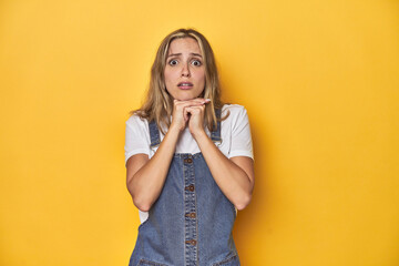 Wall Mural - Young blonde Caucasian woman in denim overalls posing on a yellow background, praying for luck, amazed and opening mouth looking to front.