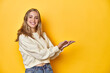 © Asier - Young blonde Caucasian woman in a white sweatshirt on a yellow studio background, holding a copy space on a palm.